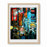 A framed art print depicting a vibrant, crowded Shinjuku street at night with glowing neon signs in red, blue, and yellow, set in a light oak wood frame with a white mat.