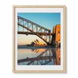 A framed photographic art print of Sydney Harbour, featuring the Sydney Opera House, the Harbour Bridge, and warm sunset reflections on the water, presented in a light natural wood frame with a white mat.