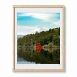 Framed wall art print showing a scenic landscape with a snow-capped mountain, green forest, calm water, and a bright red torii gate reflected in the lake. The print is displayed in a light natural wood frame with a white mat against a clean white background.