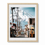 An oak framed photographic art print of a Japanese street scene with a snow-covered mount fuji in the background, shown against a plain white wall.