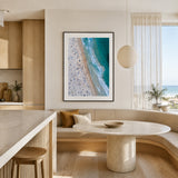 A framed vertical art print of an aerial bondi beach scene with turquoise waves and a sandy shore, displayed on a beige wall in a modern, sunlit kitchen and dining area.