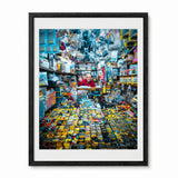A framed photographic print of a crowded, colorful toy and hobby shop interior in Japan, featuring a person (akihabara man) behind a counter surrounded by shelves and bins of small items. The print is presented with a white mat and a black frame against a plain white background.