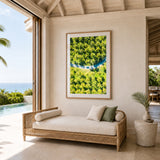 A large framed art print featuring a lush green tropical forest canopy with a blue river, displayed on a beige wall in a modern, sunlit villa interior above a wicker daybed.