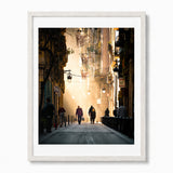 A framed photographic art print of a narrow Barcelona street with warm golden light, hanging lamps, and silhouetted pedestrians, shown in a white frame.