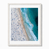 A framed aerial photograph of a bondi beach scene, featuring a sandy shore crowded with small figures, turquoise ocean water, and white surf, presented in a light wood frame against a white background.