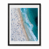 A framed aerial photograph of a bondi beach scene, showing a crowded sandy shore on the left and turquoise ocean waves on the right, presented in a black frame with a white mat against a light gray background.