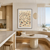 A framed vertical art print depicting a crowded bondi beach scene with tiny figures and umbrellas, hanging on a beige wall in a modern, sunlit kitchen and dining area.