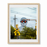 A Japan framed art print featuring a creative photograph of a giant white Buddha statue wearing black sunglasses with red lenses, set against a background of green trees and utility poles. The print is housed in a light natural wood frame with a white mat, shown against a plain white background.