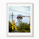 A Japan framed art print featuring a creative photograph of a giant white Buddha statue wearing black sunglasses with red lenses, set against a backdrop of green trees, utility poles, and yellow railroad crossing signals. The print is presented in a white mat and a modern white frame against a clean white background.