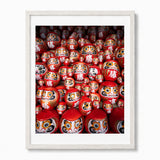 A framed photographic print of a dense arrangement of red Daruma doll figurines, presented in a white frame against a light gray background.