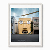 A framed art print featuring a whimsical photograph of face house in Kyoto, Japan - a yellow building with circular windows resembling a face, with a cyclist riding on the street in front. The print is framed in a white wood frame with a white mat against a white background.