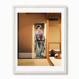 A framed art print featuring a traditional Japanese geisha in a colorful kimono standing in a tatami room, presented in a white frame with a white mat on a light gray background.