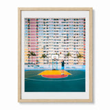 A framed photographic art print depicting a person playing basketball in a hong kong colorful high-rise building, presented in a light wood frame with a white mat.
