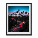 A framed vertical art print featuring a long-exposure photograph of a palm tree-lined Los Angles city street at dusk, with red light trails from cars, a blue and pink sky, and a white airplane contrail. The print is matted in white and enclosed in a black frame, shown against a plain white background.