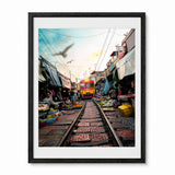A framed photographic art print depicting a vibrant railway market scene in Thailand with a yellow train on tracks, surrounded by market stalls. The print is presented in a white mat and a thick black wooden frame against a clean white background.