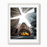 A framed photographic art print of a New York City street scene, featuring a yellow cab and surrounding skyscrapers viewed from a low-angle perspective, set in a white frame against a light gray wall.