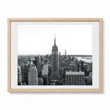 Framed black and white New York City skyline art print featuring the Empire State Building, shown in a light natural wood frame with a white mat on a white background.