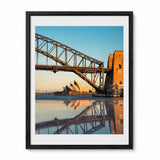 Framed wall art print showing a scenic photograph of the Sydney Harbour Bridge and Opera House reflected in calm water, with a black frame and white mat on a light gray background.