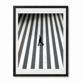A framed black and white street art print depicting a man walking across a zebra crossing, presented in a black frame with a white mat against a light gray background.