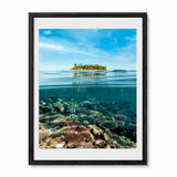 A framed photographic art print depicting a split view of a tropical island above the waterline and a clear underwater reef scene below, presented in a black frame with a white mat against a plain white background.