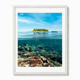 A framed split-level photograph of a tropical island above the waterline and coral reef below, presented in a white frame against a light gray background.