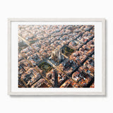 A framed aerial photograph of Barcelona's cityscape, featuring the Sagrada Ilesia at the center, surrounded by dense historic buildings and streets, presented in a light-colored wooden frame with a white mat against a white background.