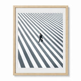 Framed wall art featuring a high-angle black and white photograph of a man walking across a striped crosswalk, presented in a wood frame with a white mat against a light gray background.