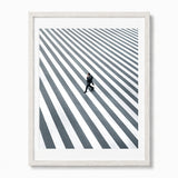 Framed wall art featuring a high-angle black and white photograph of a man walking across a striped crosswalk, presented in a white frame with a white mat against a light gray background.
