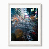 A framed art print depicting a high-angle night view of Shibuya Crossing in Japan featuring vibrant neon signs, pedestrians, and street traffic. The print is housed in a thick white wooden frame and is displayed against a clean white wall.