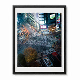 A framed art print depicting a high-angle view of a busy Shibuya Crossing Japan at night, featuring vibrant neon signs and pedestrians. The print is framed in a black frame with a white mat, set against a plain white background.
