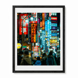 A framed art print depicting a vibrant, neon-lit street scene in Sinjuku, Japan at night, featuring colorful signs and silhouettes of pedestrians. The print is housed in a black frame with a white mat, shown against a plain white background.