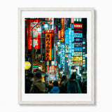 A framed art print depicting a vibrant, crowded Shinjuku Japan at night with glowing neon signs in red, blue, and yellow, set in a white wood frame with a white mat.