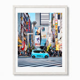 A framed photographic art print of a vibrant street scene in Tokyo, Japan, featuring a light blue compact car on a black-and-white striped crosswalk surrounded by tall buildings and colorful neon signs. The print is housed in a white frame with a white mat, set against a clean white background.