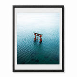 Framed wall art featuring an aerial photograph of a red torii gate in Japan standing in calm blue-green water, presented in a black frame with a white mat against a light gray background.