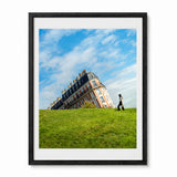 A framed art print featuring the sinking of house of Montmartre in Parisg on a grassy hill under a blue sky with white clouds, with a person walking on the grass. The print is framed in a black frame with a white mat against a white background.