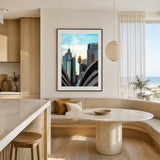 A framed vertical art print of a modern city skyline with the Sydney Tower visible, hanging on a beige wall in a contemporary, sunlit kitchen and dining area.