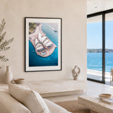A framed vertical art print of an aerial view of the Sydney Opera House in blue water, displayed on a beige wall in a modern, sunlit living room with a sofa, decorative vases, and a large window overlooking the ocean.