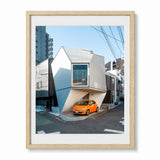 A photograph of a small house in Tokyo Japan with an orange car parked in front, presented in a wood frame with a white mat against a light gray background.