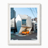 A photograph of a small house in Tokyo Japan with an orange car parked in front, presented in a white frame with a white mat against a light gray background.