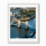 A framed aerial photograph of London's Tower Bridge and surrounding cityscape, shown against a clean white background. The print features a white frame and white mat, with the city and bridge depicted in warm golden light.