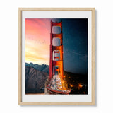 Framed wall art featuring a split view of the Golden Gate Bridge, with a sunset sky on the left and a starry night sky on the right, shown in a light wood frame against a white background.