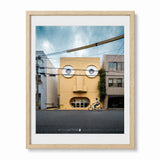 A framed art print featuring a whimsical photograph of face house in Kyoto, Japan - a yellow building with circular windows resembling a face, with a cyclist riding on the street in front. The print is framed in a light natural woode with a white mat against a white background.