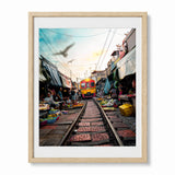 A framed photographic art print depicting a vibrant railway market scene in Thailand with a yellow train on tracks, surrounded by market stalls. The print is presented in a white mat and a light wood frame against a clean white background.