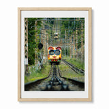 A framed photographic print of a colorful Ikoma Cable Car in Japan, on railway tracks surrounded by green trees and overhead power lines, presented in a light wood frame with a white mat against a white background.