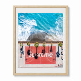 A framed art print featuring an aerial photograph of a red basketball court with palm trees and the word 'Supreme' overlaid, at a California beach. Set in a light wood frame with a white mat.