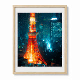 A framed art print of the Tokyo Tower illuminated in vibrant orange-red tones against a dark blue and teal city skyline at night, presented in a light wood frame with a white mat.