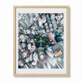 Framed aerial photograph of a dense Hong Kong urban cityscape with a central green park and airplane, shown in a light wood frame with a white mat against a white background.