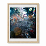 A framed art print depicting a high-angle night view of Shibuya Crossing in Japan with illuminated billboards and streetlights, presented in a light natural wood frame with a white mat against a white background.