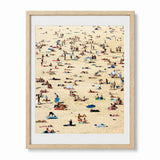 A framed art print depicting a crowded Bondi beach scene with many small figures, towels, and umbrellas on a sandy beige background, shown in a light natural wood frame with a white mat.