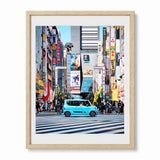 A framed art print depicting a vibrant street scene in Tokyo, Japan, featuring a light blue car crossing a zebra crosswalk amidst towering buildings and colorful neon signs. The print is presented in a light natural wood frame with a white mat, set against a clean white background.